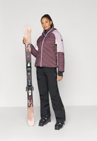 Smučarska oprema z žensko, ki drži večbarvno izolirano jakno, črne smučarske hlače in smuči z nežnim prelivom ter vezmi.