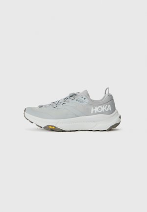 Light gray athletic shoe with a mesh upper, reinforced toe, and rubber Vibram sole. Features toggle lacing and HOKA branding on the side.