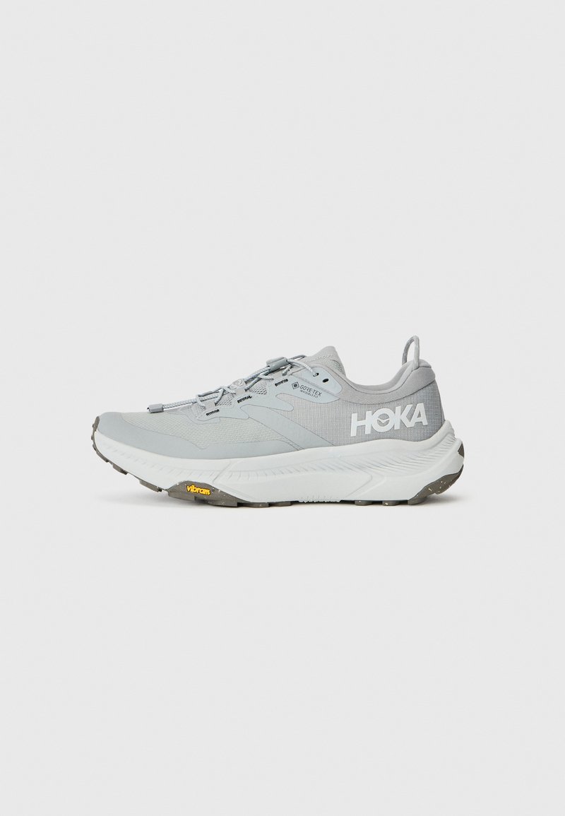 Sapatilha atlética cinza claro com parte superior em malha, biqueira reforçada e sola de borracha Vibram. Apresenta atacadores de pressão e a marca HOKA na lateral.