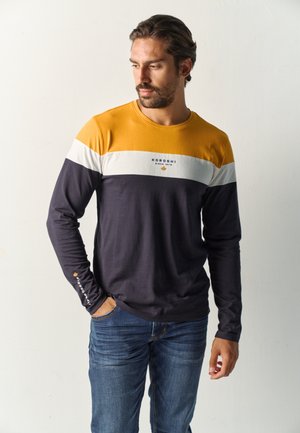Hombre con barba que lleva una camisa de manga larga combinada en colores mostaza, blanco y azul marino con vaqueros azules, de pie con una mano en el bolsillo.
