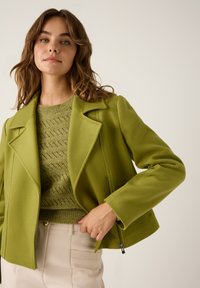 Grüne Jacke mit strukturiertem Kragen und Reißverschluss-Taschen, über einem strukturierten grünen Strickpullover getragen, kombiniert mit hellen Hosen.