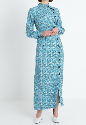 Frau trägt ein blaues, langärmeliges, knöchellanges Kleid mit Blumenmuster und schwarzen Knöpfen auf der Vorderseite sowie weiße Pumps; sie steht vor einer weißen Wand.