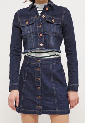 Kvinde iført en mørkeblå cropped denimjakke med knaplukning og et matchende denimnederdel med knapper foran.