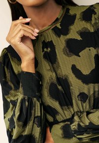 Blouse texturée verte et noire avec un motif léopard, présentant un col rond et des manches bouffantes, détaillée avec une taille froncée et des poignets boutonnés.