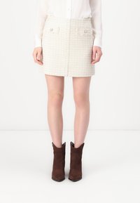 Witte tweed mini-rok met een gestructureerd patroon, voorzien van twee voorkeurszakken en decoratieve knopen. Draag het met bruine suède enkellaarsjes.