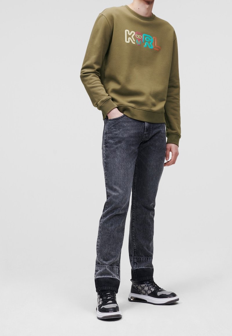 Personne portant un sweat-shirt vert olive avec un logo "KRL" coloré, un jean noir délavé à l'acide, et des baskets noir et blanc, debout devant un fond uni.