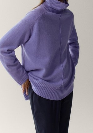 Person wearing a loose lavender turtleneck sweater with ribbed cuffs and hem, paired with black pants, against a plain background.