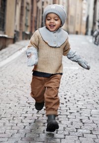 Hellbeige gestrickter Pullover, grauer Kragen, graue Handschuhe, braune Cordhosen, grüne Stiefel; Kind läuft auf einer Kopfsteinpflasterstraße.