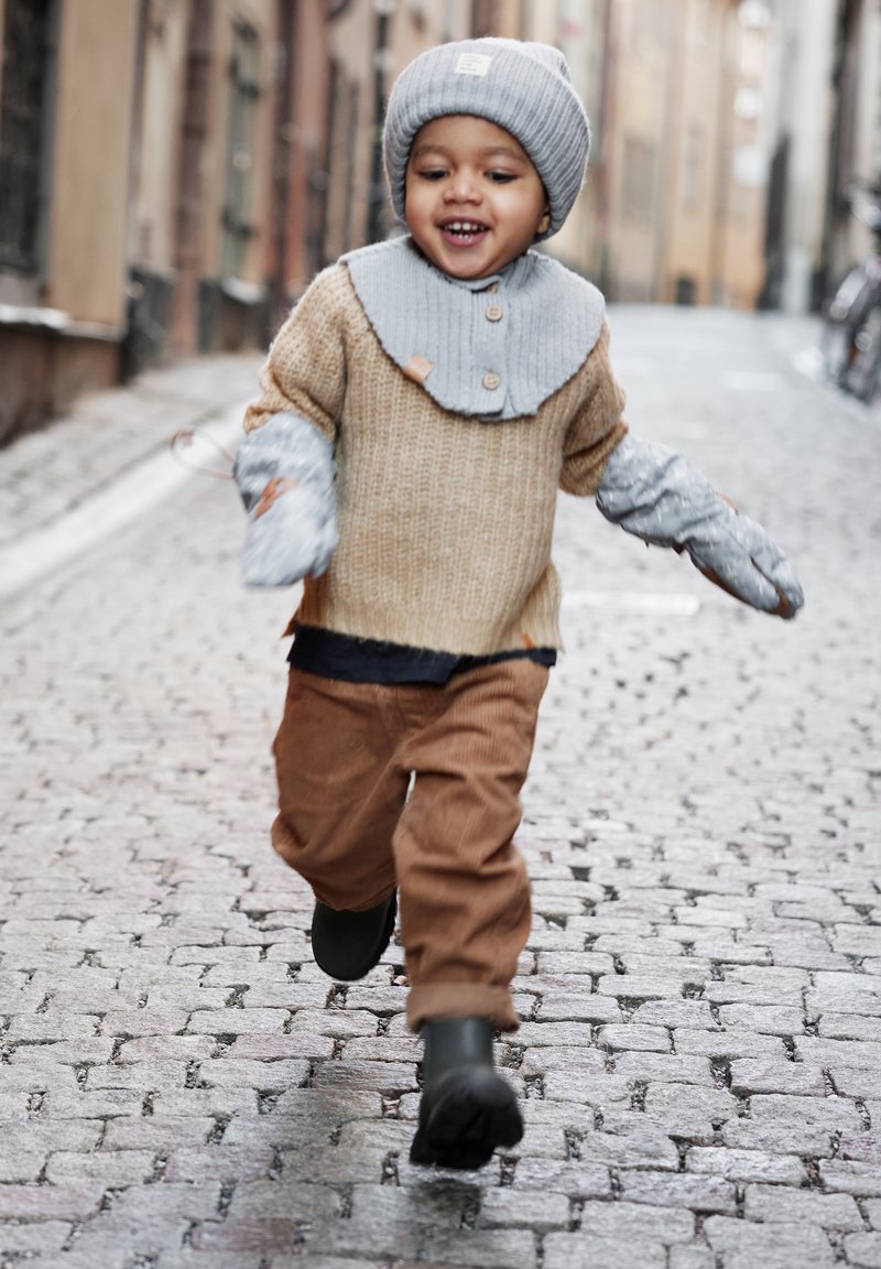 Hellbeige gestrickter Pullover, grauer Kragen, graue Handschuhe, braune Cordhosen, grüne Stiefel; Kind läuft auf einer Kopfsteinpflasterstraße.
