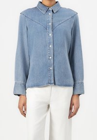 Personne portant une chemise en denim bleu clair avec des boutons blancs et des manches larges retroussées, associée à un pantalon blanc.