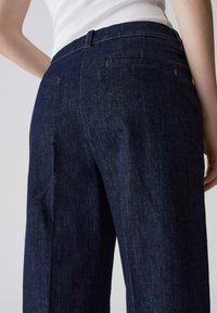 Pantalons en denim foncé avec taille haute, présentant un design épuré, des poches arrière et des coutures contrastées pour plus de détails.