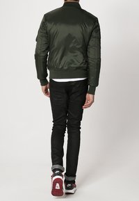 Veste bomber vert olive avec une texture lisse, des poignets et un ourlet côtelés. Portée sur une chemise blanche, associée à un jean noir et des baskets.