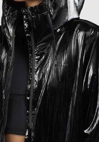 Veste noire brillante à capuche avec fermeture éclair et cordons portée sur un top noir, montrant le haut du torse et le cou.