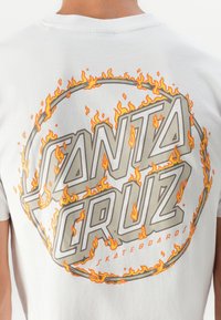 T-shirt branca de algodão com um gráfico circular com "SANTA CRUZ" em letras maiúsculas e negrito, rodeado por designs de chamas em laranja e amarelo.
