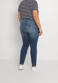 Person som bär mid-rise slim-fit blå jeans, en svart och vit randig t-shirt och vita Nike sneakers, stående mot en vit vägg.