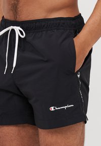 Juodi maudymosi šortai, pagaminti iš lengvos medžiagos, su raišteliu liemenyje, šoninėmis kišenėmis ir balta Champion logotipo detale ant kojos.