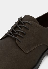 Σκούρο καφέ παπούτσι από σουέντ με σχέδιο με κορδόνια, διαθέτοντας ανάγλυφο άνω μέρος, στρογγυλεμένη μύτη και διακριτικό ράψιμο κατά μήκος της άκρης.