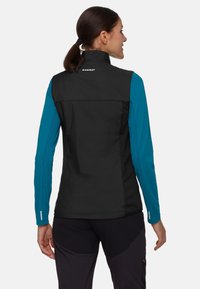 Frau mit dunklem Pferdeschwanz, die eine schwarze Mammut-Weste über einem türkisfarbenen Langarmshirt und schwarzen Hosen trägt, von hinten vor einem weißen Hintergrund.