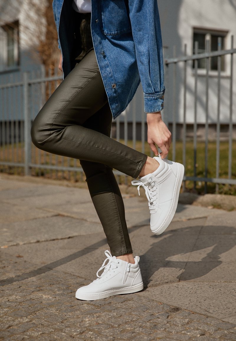 Person in olivgrünen Hosen und einer Jeansjacke, der weiße High-Top-Schuhe auf dem gepflasterten Gehweg in der Nähe eines Metallzauns anpasst.