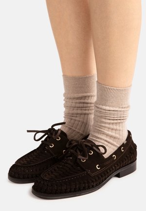 Jambes portant des chaussettes beiges côtelées et des chaussures à lacets en cuir tissé noir avec des œillets dorés sur un fond clair.