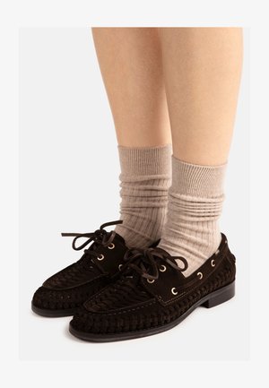 Jambes portant des chaussettes beiges côtelées et des chaussures à lacets en cuir tissé noir avec des œillets dorés sur un fond clair.