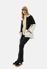 Chaqueta en bloques de color negro y crema, suéter texturizado beige, jeans de campana negros y zapatillas negras con suelas marrones; gorra beige.