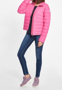 Gevoerde roze jas met een ritssluiting aan de voorkant, een staande kraag en een gevoerd ontwerp; gecombineerd met blauwe skinny jeans en lichtpaarse sneakers.