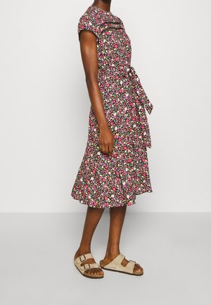 Femme portant une robe fleurie à manches courtes et longueur au genou, avec une ceinture à la taille, associée à des sandales beiges à double bride, se tenant de profil sur un fond uni.