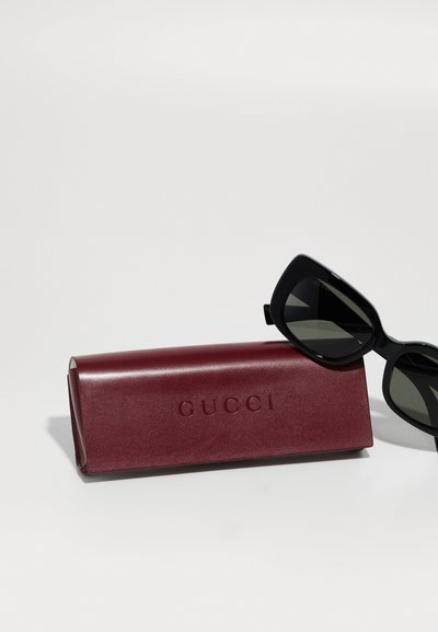 Mustat aurinkolasit, joissa on gradienttipinnat, lepäävät burgundin värisessä nahkakotelossa, jossa on syvävaikutteinen "GUCCI"-logo. Kotelo on sileäpintainen ja rakenteeltaan selkeä.