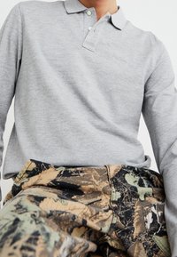 Harmaa pitkähihaisella polo paidalla, jossa on kaulus ja kaksi nappia, yhdistettynä camo-kuosisesti housujen kanssa, joissa on vihreitä, ruskeita ja mustia lehtikuvioita.