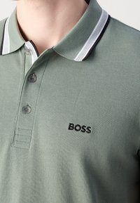 Gros plan d'un polo vert clair avec un col rayé blanc et noir et un logo "BOSS" noir brodé sur le côté gauche de la poitrine.