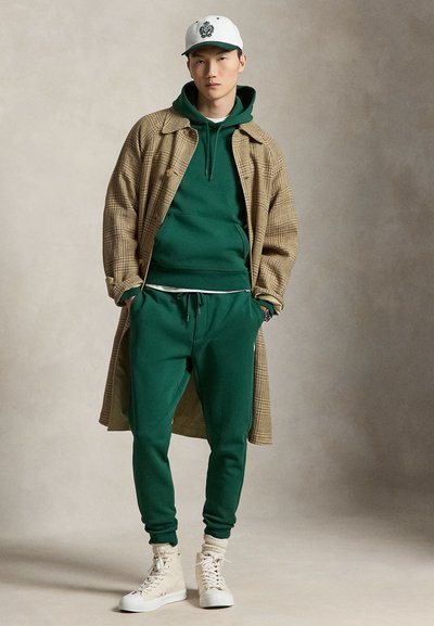 Jonge man draagt een groene hoodie en joggingbroek, een beige geruite lange jas, witte hoge sneakers en een witte pet met een zwart embleem.