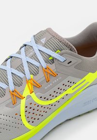 Chaussure de trail Nike en mesh gris avec des lacets bleus, des accents orange et des détails jaune néon. Présente des perforations pour la respirabilité.
