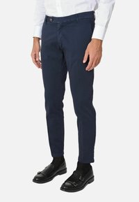 Chino blu marina in cotone con vestibilità slim, dotati di tasche standard, chiusura con bottone, indossati con mocassini neri e calze scure.