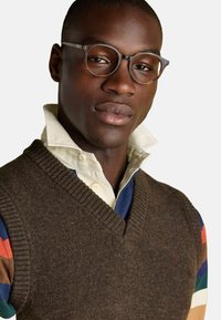 Pruun kudumine vest, millel on V-kaelus, kantakse kraega särgi peal. Särgil on mitmevärvilised triibulised varrukad. Ümmarguste raamidega prillid.