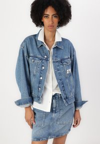 Τζάκετ από denim σε ανοιχτό μπλε χρώμα με ασημένια κουμπιά, φθαρμένες λεπτομέρειες και δύο μπροστινές τσέπες, συνδυασμένο με λευκό πουκάμισο με γιακά και φούστα από denim.