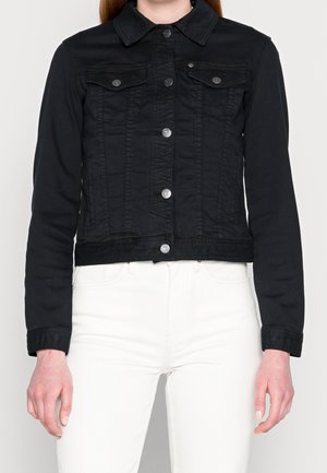 Kvinde iført en tætsiddende sort denimjakke med sølvknapper over hvide bukser, stående mod en ensfarvet lys baggrund.