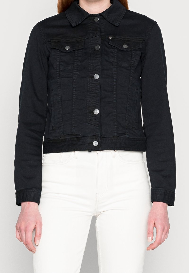 Femme portant une veste en jean noire cintrée avec des boutons argentés sur un pantalon blanc, debout devant un fond clair uni.