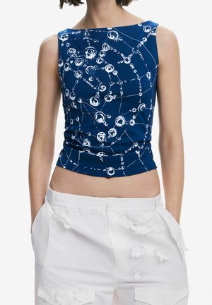 Kobieta w niebieskim topie bez rękawów z białymi abstrakcyjnymi wzorami w kształcie bąbelków oraz w wysokich białych szortach z kwiatowymi aplikacjami.