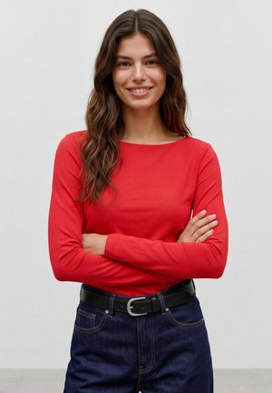 Femme aux longs cheveux bruns ondulés portant un haut rouge à manches longues et un jean bleu foncé avec une ceinture noire, souriante, les bras croisés.
