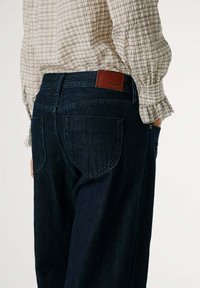 Jeans de mezclilla azul oscuro con un parche de cuero en la cinturilla trasera. Presentan dos bolsillos traseros y un diseño de pierna recta.