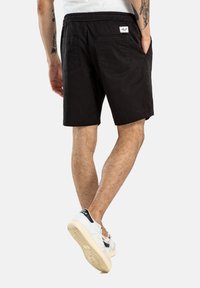 Pantalones cortos negros con cintura elástica, dos bolsillos traseros y una pequeña etiqueta con el logo. La tela parece ligera y casual.