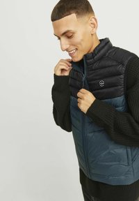 Vest cu umplutură în nuanța bleumarin închis și negru, cu un design prostu numit "quilted", guler în picioare și închidere cu fermoar. Mâneci ribbate negre oferă textură.