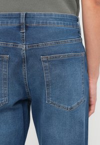 Κοντινή λήψη πίσω μέρους μπλε τζιν με ορατές τσέπες και ραφές, φορεμένο με γκρι υφασμάτινο πουκάμισο.