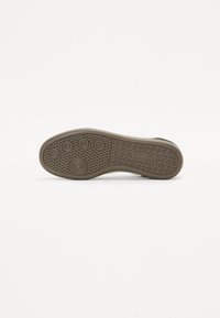 Suola di scarpa in gomma con una superficie strutturata e decorata, caratterizzata da impugnature circolari e branding. Il colore è marrone chiaro con una finitura liscia.