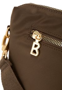 Bolso cruzado de tela marrón con un cierre dorado que tiene un tirador en forma de letra "B", superficie texturizada y correa ajustable.