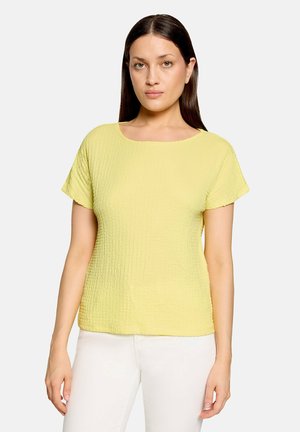Femme aux longs cheveux foncés portant un haut jaune texturé à manches courtes et un pantalon blanc, faisant face vers l'avant avec une expression neutre.