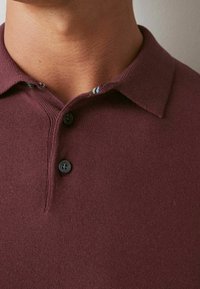 Polo bordeaux à texture côtelée, avec deux boutons noirs et un détail de col à motif présentant des rayures bleues et vertes.