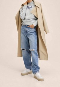 Grå sweatshirt med teksten "Touché", beige trenchcoat, lyseblå slidte jeans og hvide sneakers med beige såler.