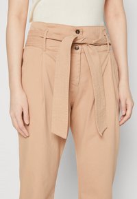 Beige hochgeschnittene Hose mit texturiertem Gürtel. Sie verfügt über einen Knopfverschluss, eine gefältete Vorderseite und seitliche Taschen für mehr Funktionalität.